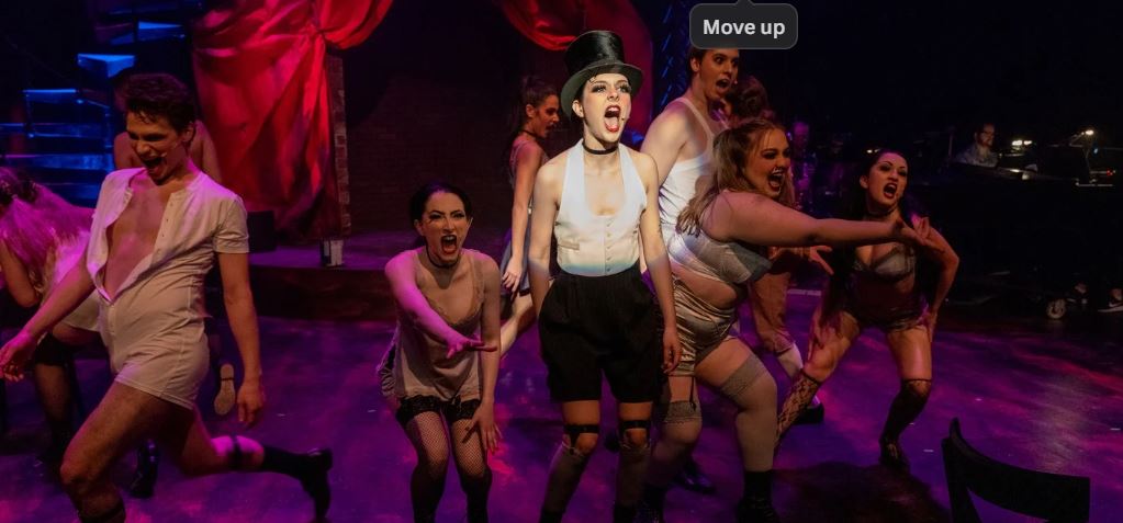 A cabaret MC stands center surrounded by scantly clad dancers.