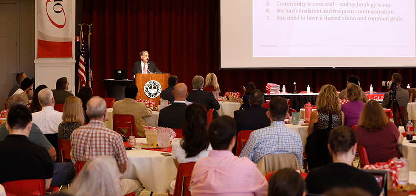 President Erik J. Bitterbaum speaks at a podium in Corey Union Function Room with faculty and staff members in attendance.