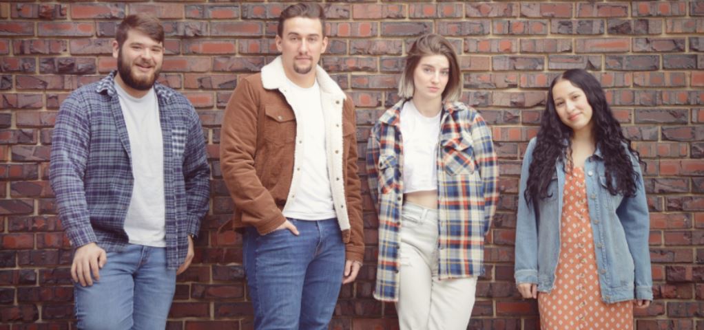 Four casually dressed young adults lean against a brick wall.