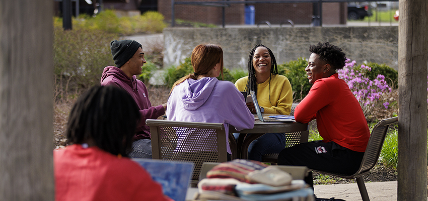 Group of four students chatting at Newmark Pavilion with Memorial Library in the background