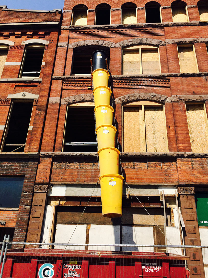 A brick building facade with a trash chute leading from an open window into a dumpster at ground level