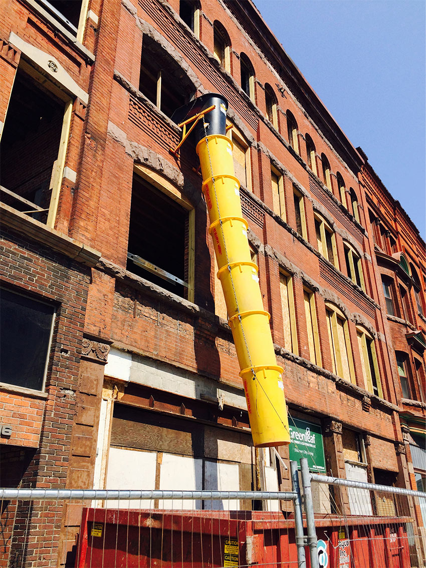 A brick building facade with a trash chute leading from an open window into a dumpster at ground level